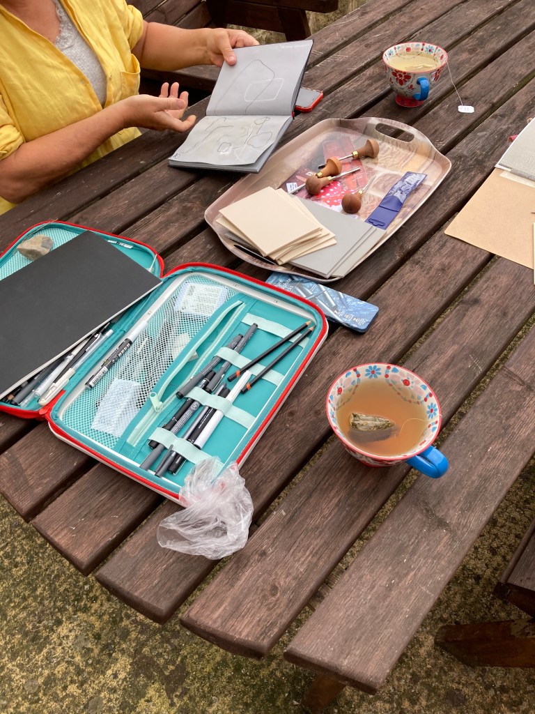 Drawing and printing materials, plus cups of tea set out on a table, one pair f hands with sketchbook