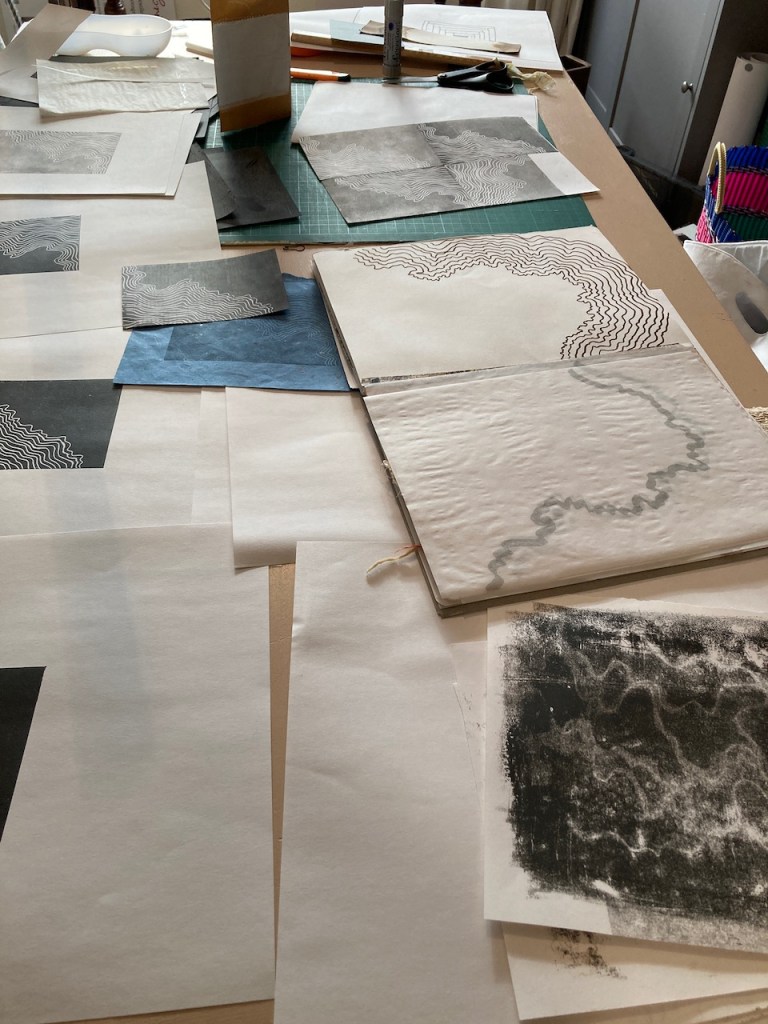 Black and white artist prints laid on a table. Prints are wiggly lines travelling across the print plate. 